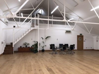A spacious modern office, once an old recording studio, with wooden floors, white walls, exposed beams, a staircase to a loft area, a meeting table with black chairs, plants, and a cabinet against the wall.