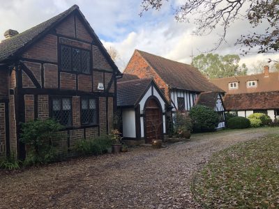 A rustic half-timbered house, reminiscent of an Old Barn Surrey, with leaded glass windows and a red brick roof sits beside a curved gravel path, surrounded by potted plants, shrubs, and trees under a partly cloudy sky.