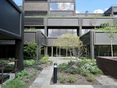 A modern building with large glass windows is surrounded by landscaped gardens, stone paths, and young trees under a partly cloudy sky. A black bollard light stands on the pathway in the foreground.