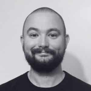 A man with a shaved head and full beard, who appears to be a Case Study Specialist, smiles slightly at the camera. He is wearing a dark shirt against a plain, light background in this black and white photo.